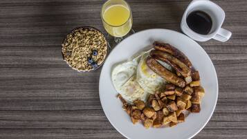 Un plato de desayuno con huevos, salchichas y papas, un tazón de granola con arándanos, un vaso de jugo de naranja y una taza de café sobre una mesa de madera.