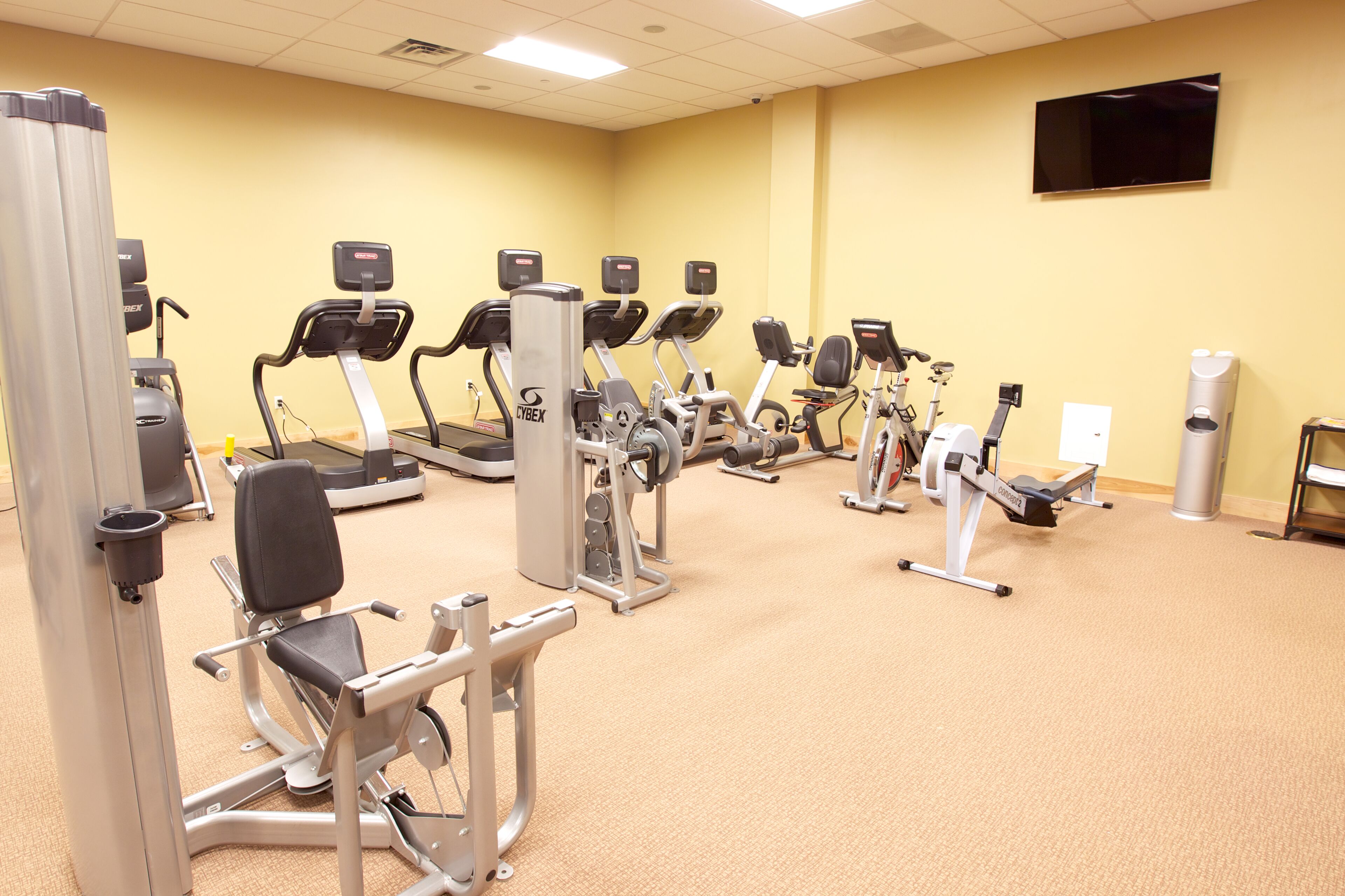 A gym with various exercise machines, a flat-screen TV on the wall, and a trash can.