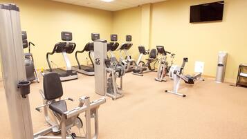 A gym with various exercise machines, a flat-screen TV on the wall, and a trash can.