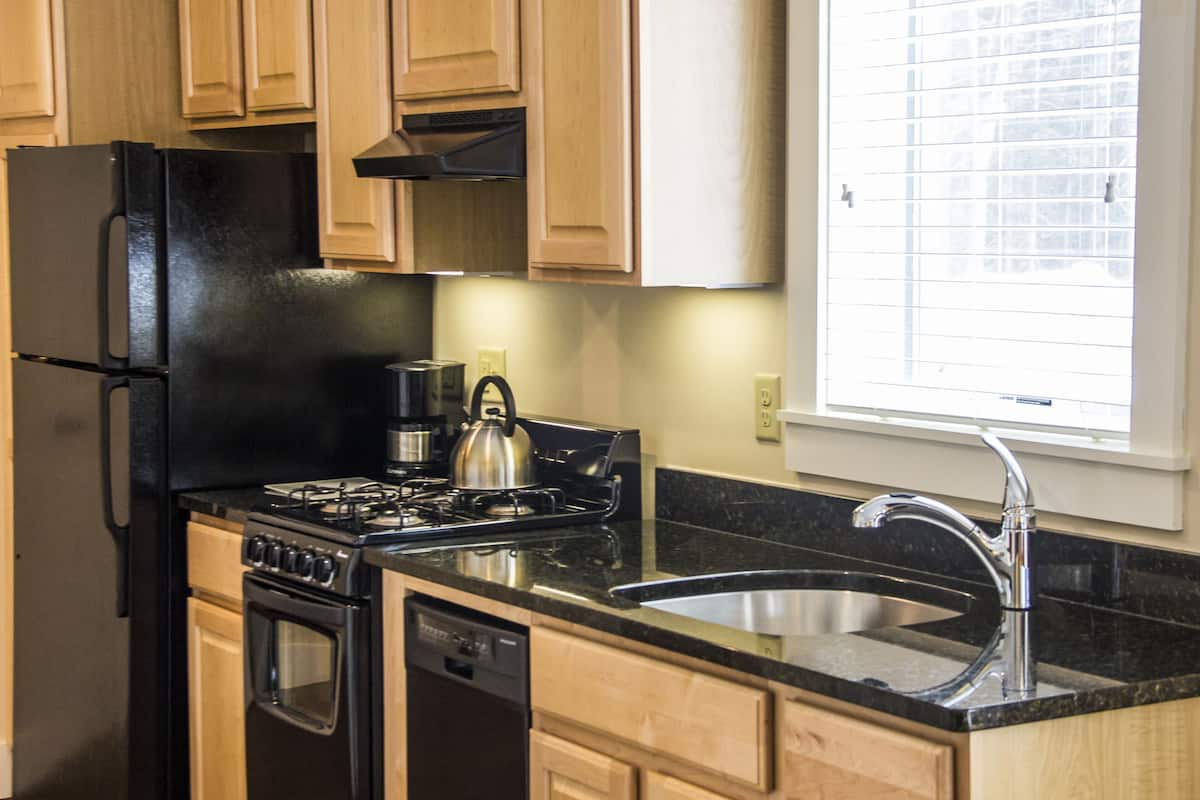 A kitchen with wooden cabinets, a black refrigerator, a stove, a kettle, and a sink.