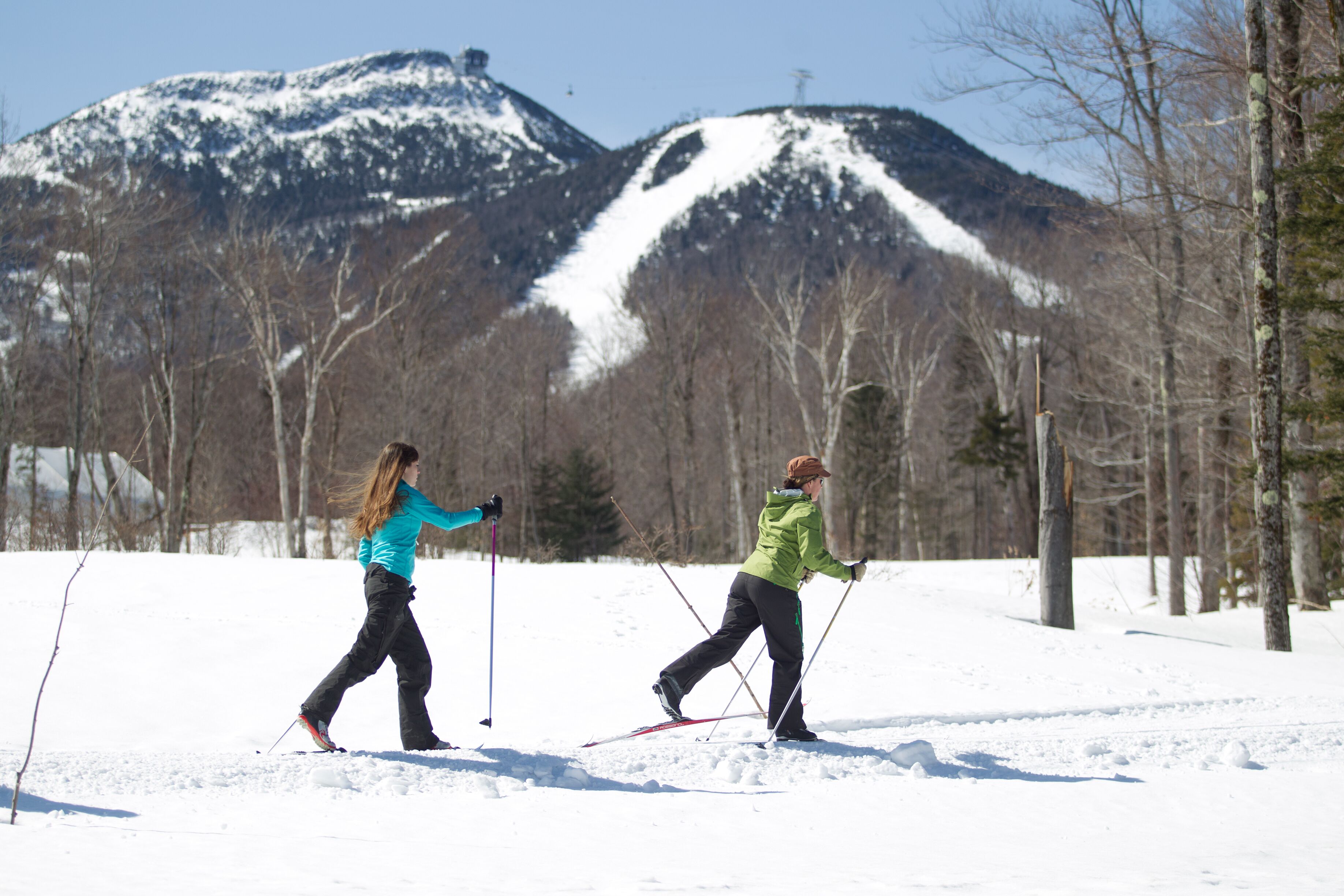 Two people cross-country skiing in a snowy landscape with mountains in the background.