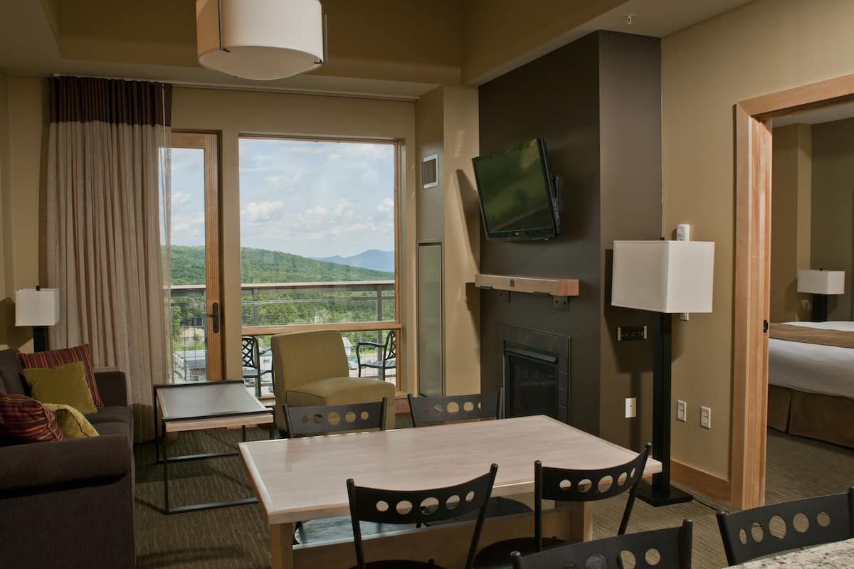 A hotel room with a dining area, a TV mounted on the wall, a fireplace, and a balcony with a scenic view.