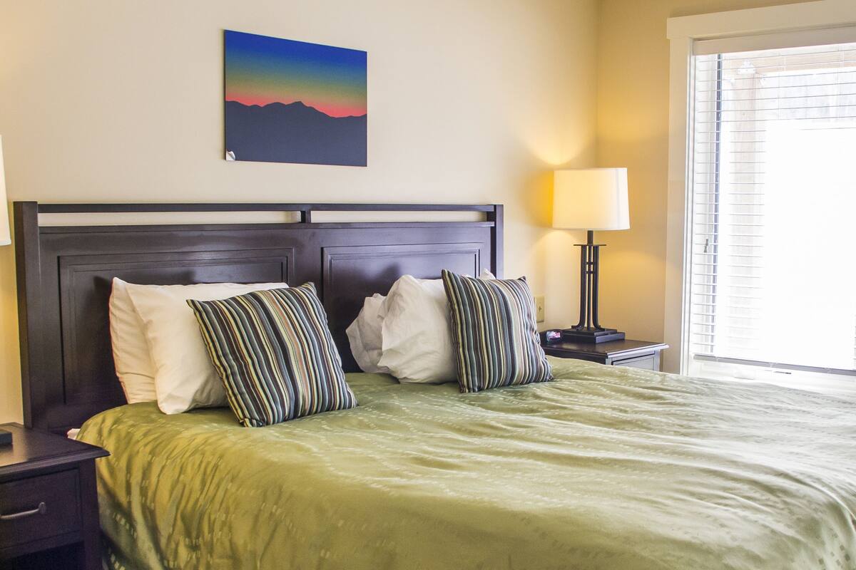 A neatly made bed with a green bedspread, two nightstands with lamps, and a painting of a sunset on the wall.