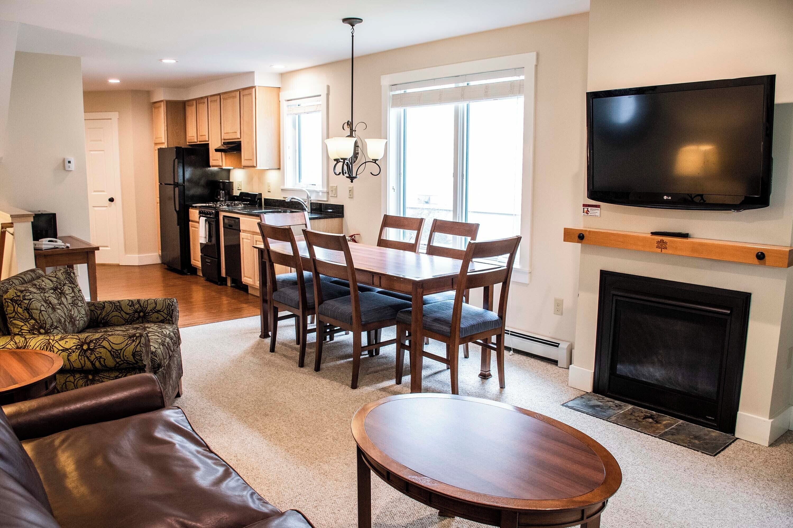 A modern living room with a fireplace, a dining area with wooden chairs, a kitchen with a black refrigerator, and a flat-screen TV mounted above the fireplace.