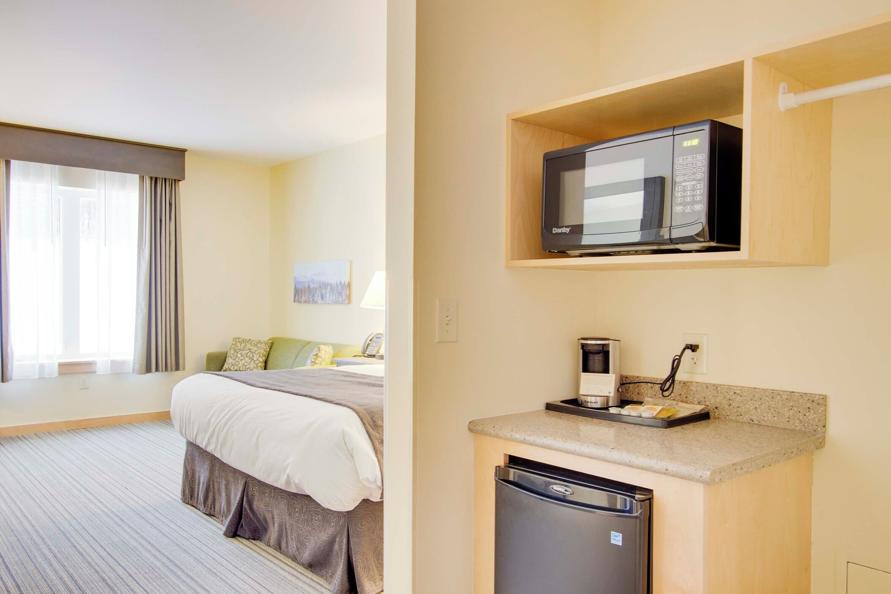 Una habitación de hotel con cama, una pequeña cocina americana equipada con microondas, cafetera y lavavajillas.