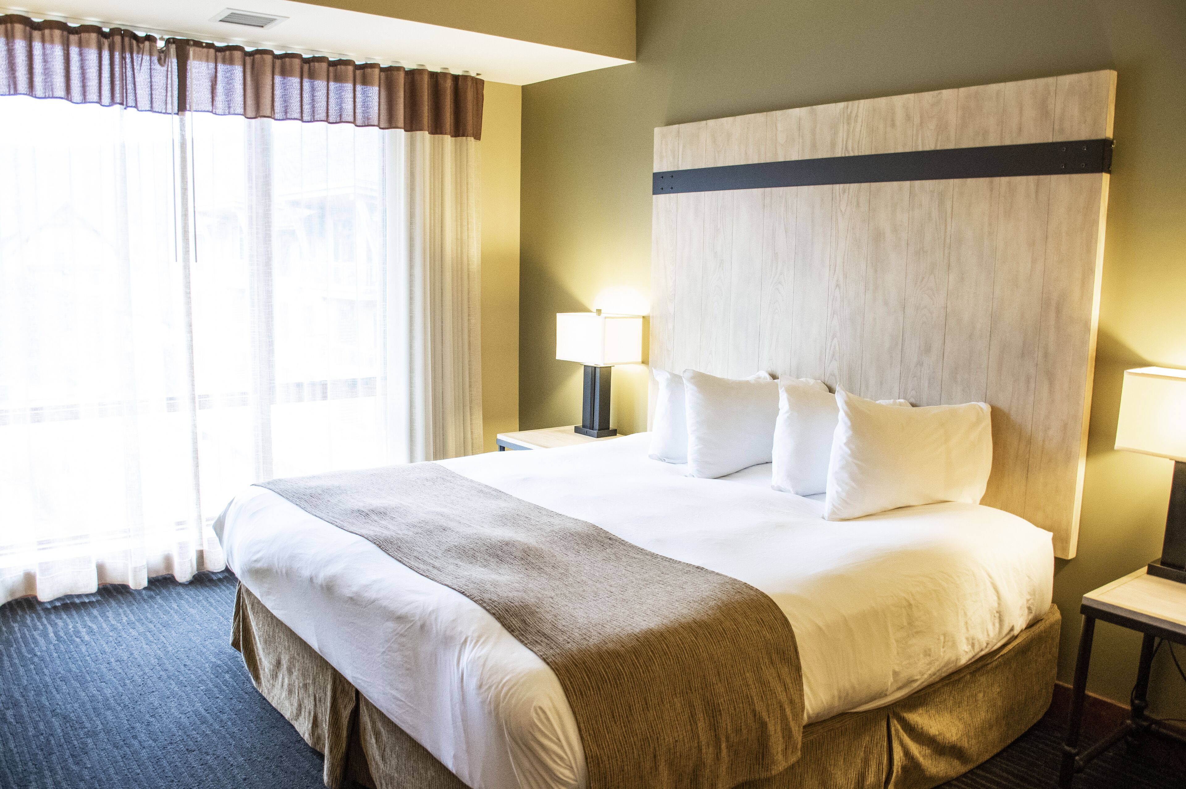 Ein Hotelzimmer mit einem großen Bett, zwei Nachttischen, einem Wandpaneel als Kopfteil und einem Fenster mit Vorhängen.