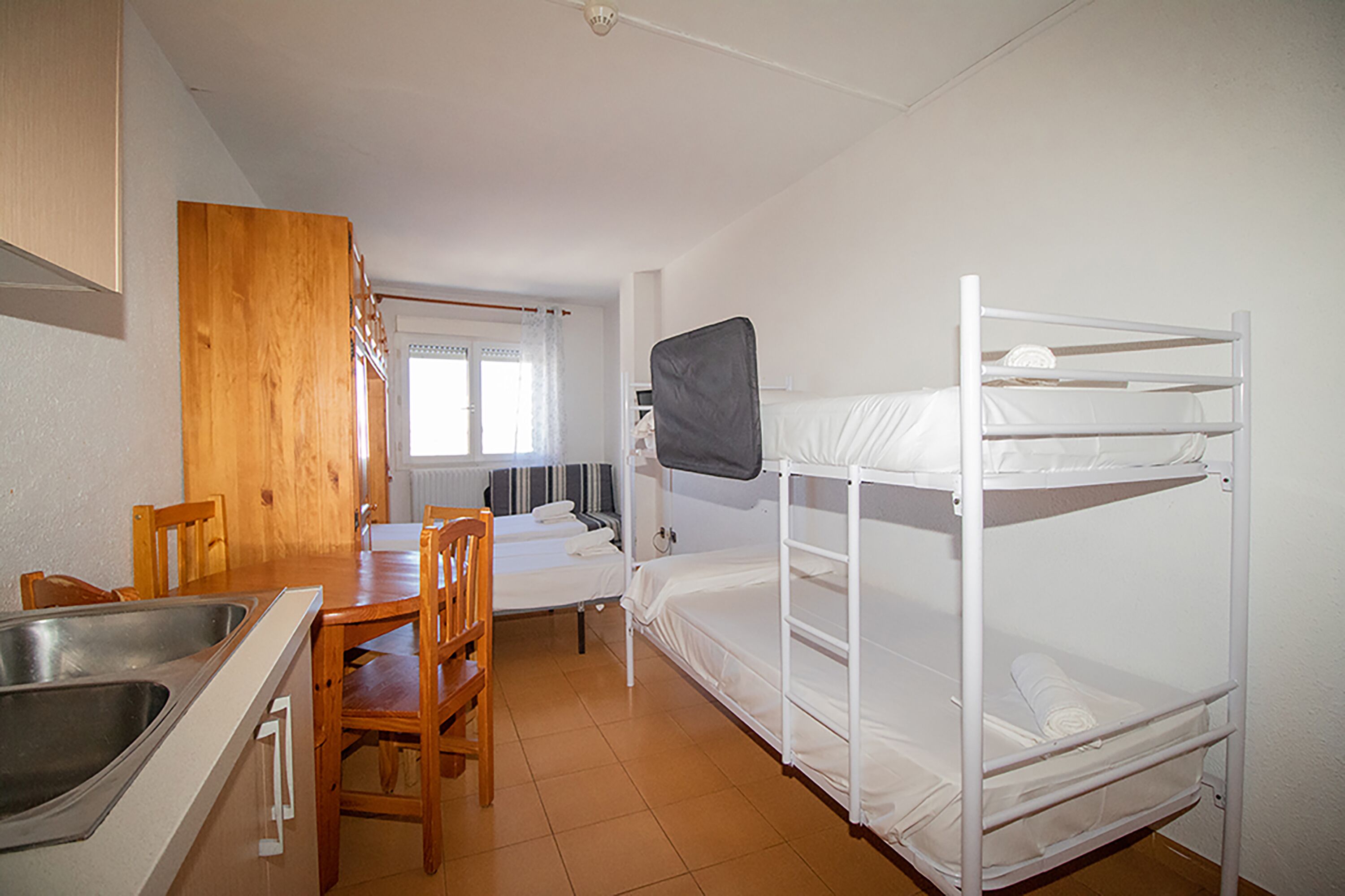 A small, compact room with a bunk bed, a wooden dining table with chairs, a kitchen area with a sink, and a window with curtains.