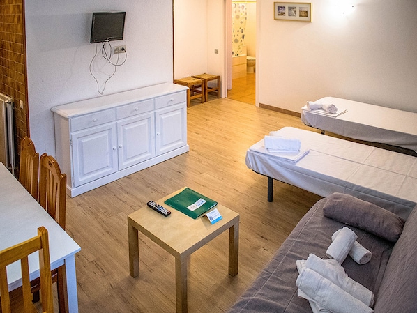 A hotel room with a wooden floor, a sofa, a small table with a remote control and a green folder, a TV mounted on the wall, and a bed with a white blanket. - Estudios Cims Pas 3000 (Pas de la Casa)