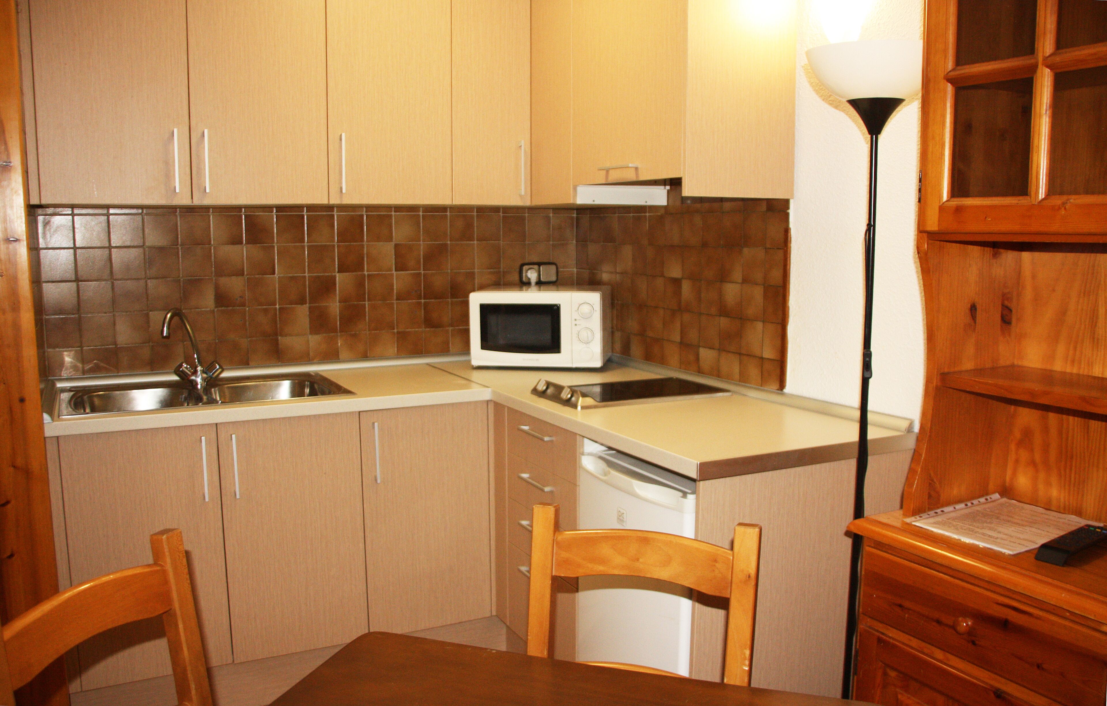 A compact kitchen with wooden cabinets, a microwave, a sink, and a dining area with chairs.