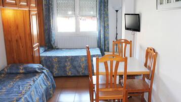 Una habitación pequeña y acogedora con una cama, una mesa de comedor con sillas, un televisor en la pared y una ventana con cortinas.