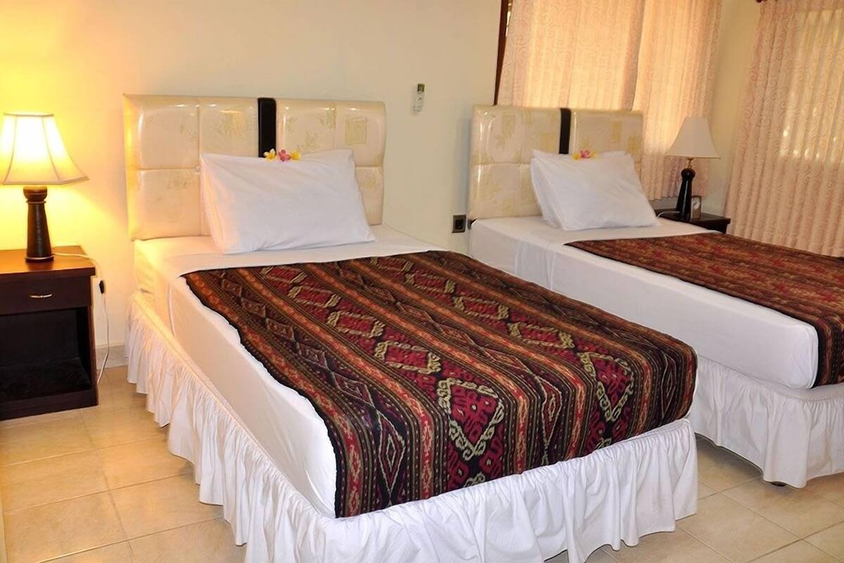 A hotel room with two beds, each with a white headboard and a patterned bedspread. There is a nightstand with a lamp on the left side of the room.