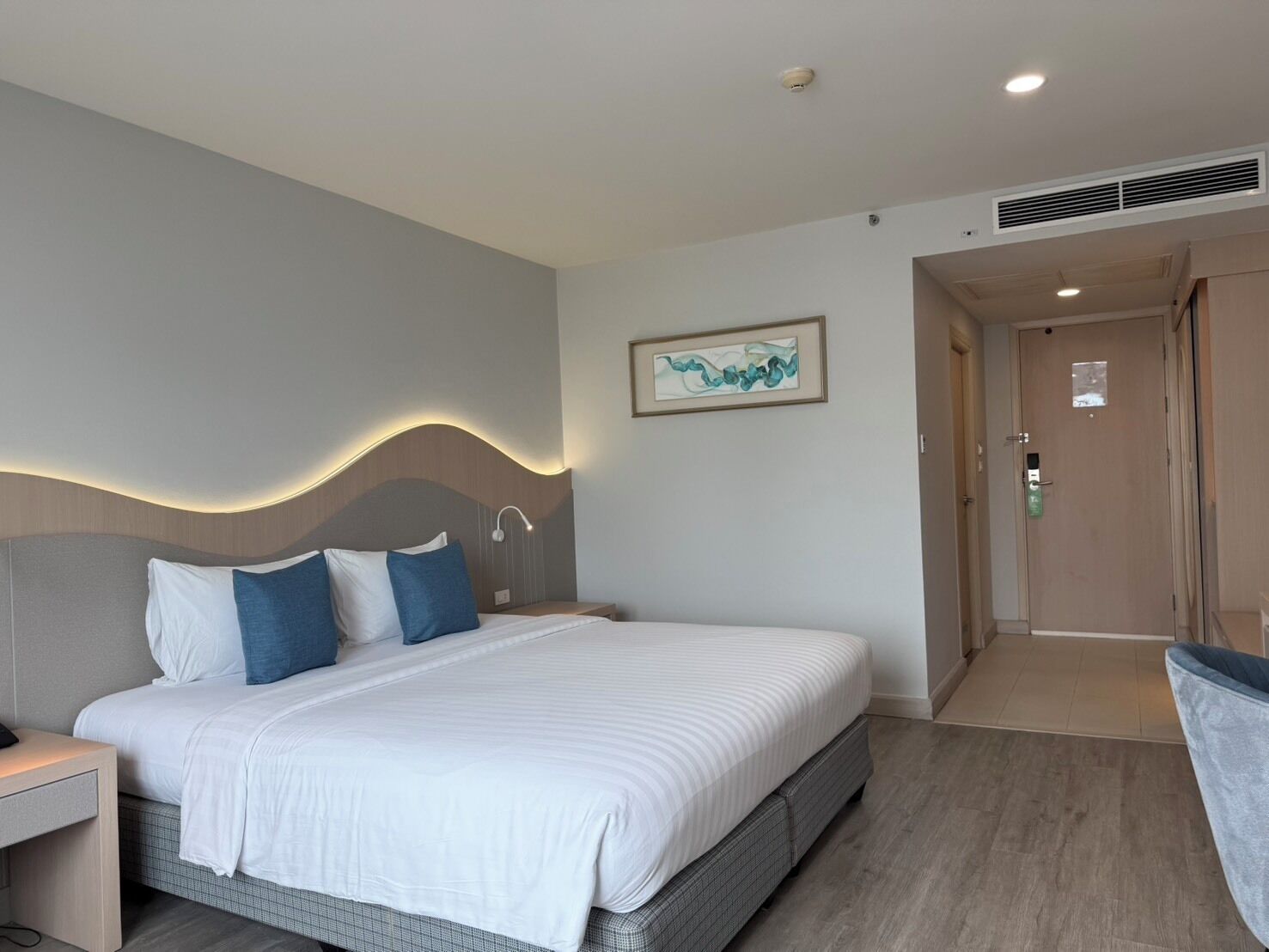 A modern hotel room with a large bed, a nightstand, a wall-mounted artwork, and a door leading to another room.