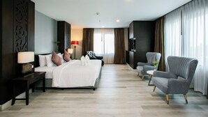 Quarto de hotel moderno com uma cama grande, área de estar com uma poltrona, TV e escrivaninha.