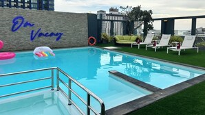 Piscina no terraço com vista clara do horizonte da cidade, parede verde com o texto 'De Férias' e cadeiras de descanso brancas ao redor da piscina.