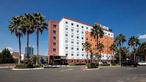A multi-story hotel with a red and white facade, palm trees, and a parking lot. - Hotel Hi! Zapopan (Zapopan)