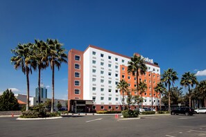 A multi-story hotel with a red and white facade, palm trees, and a parking lot. - Hotel Hi (Zapopan)