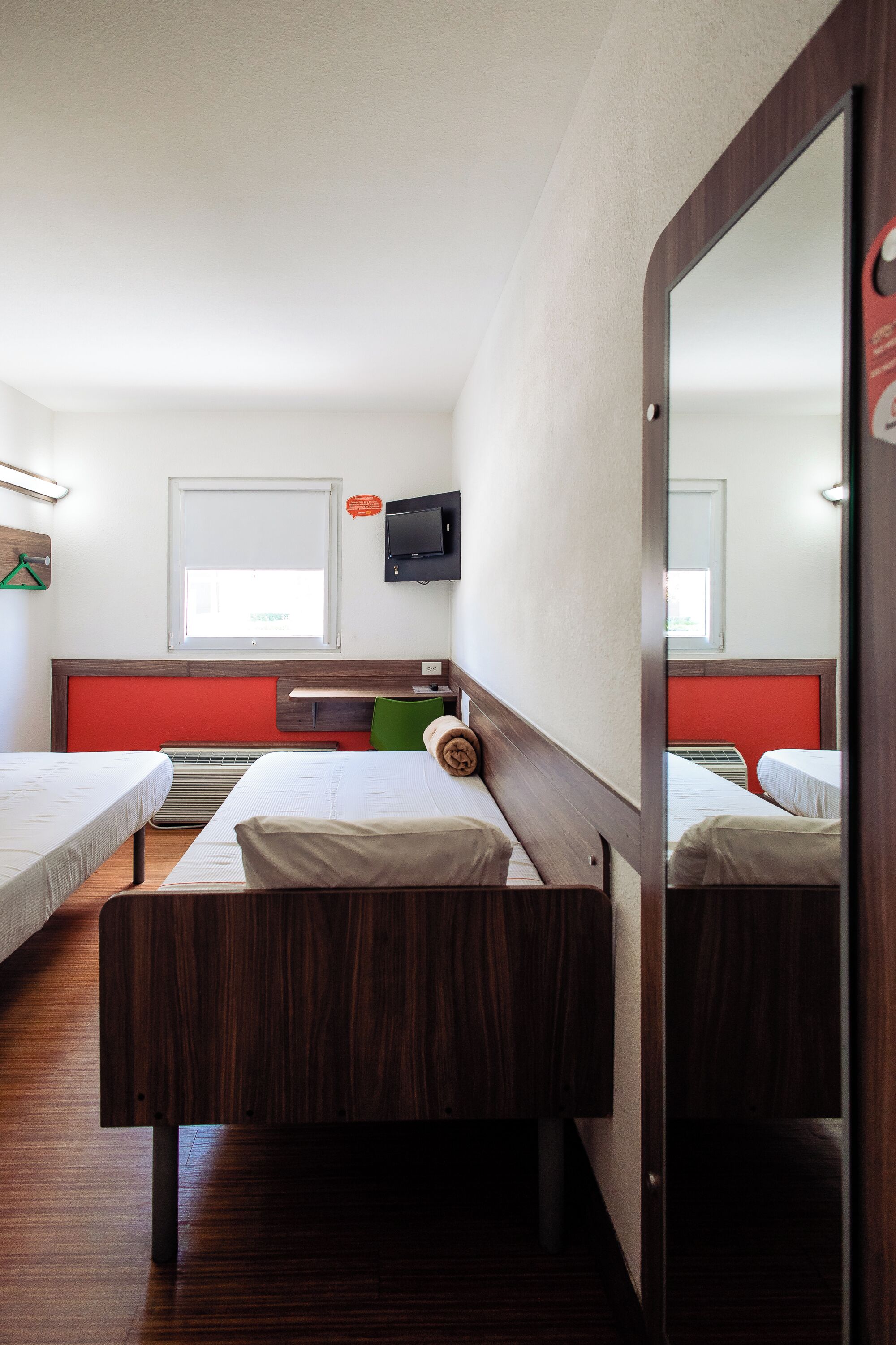 A hotel room with two single beds, a wooden headboard, a flat-screen TV, and a window with blinds.