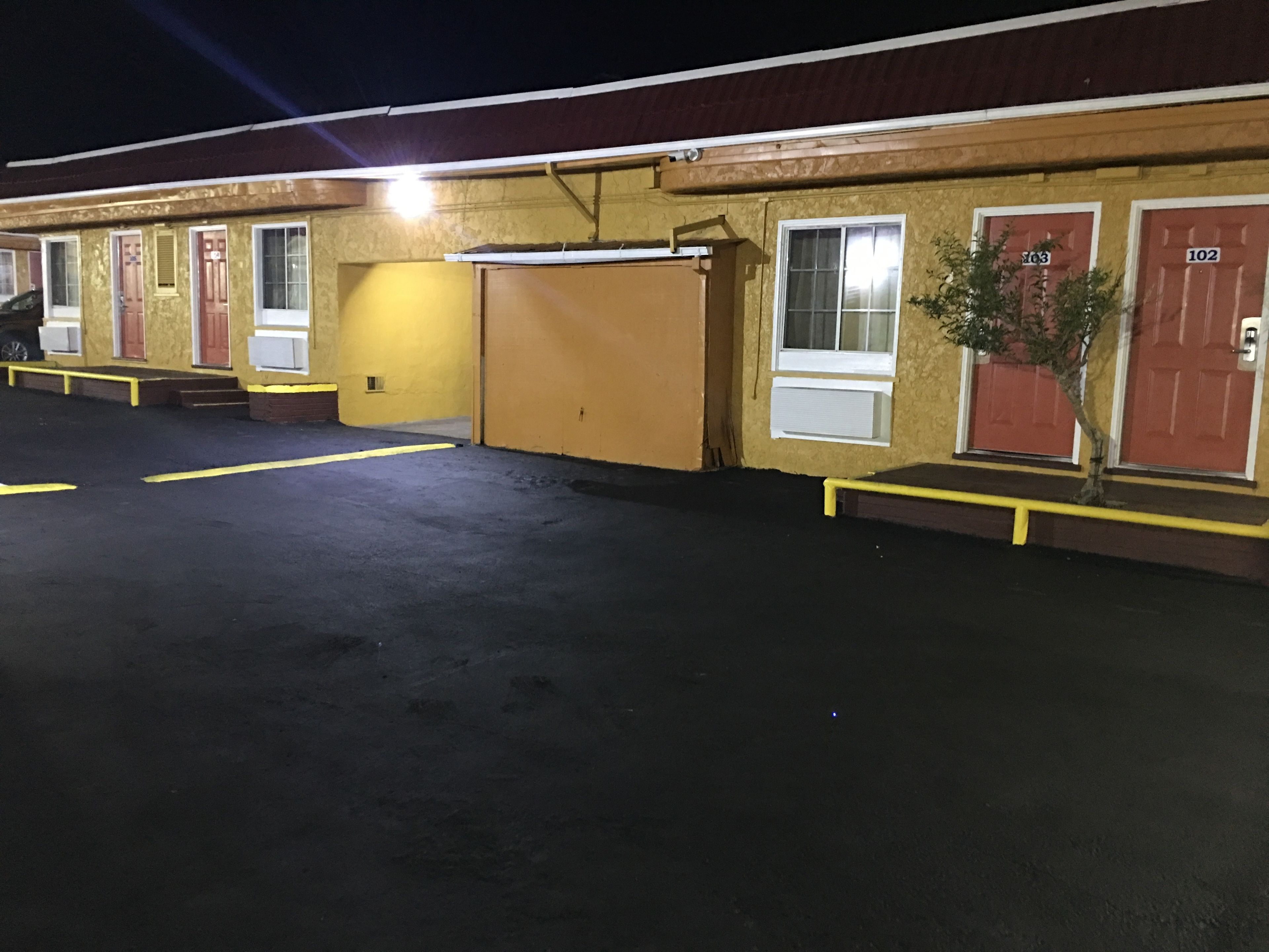 Un motel con habitaciones numeradas, una zona de estacionamiento techada y un pequeño árbol.