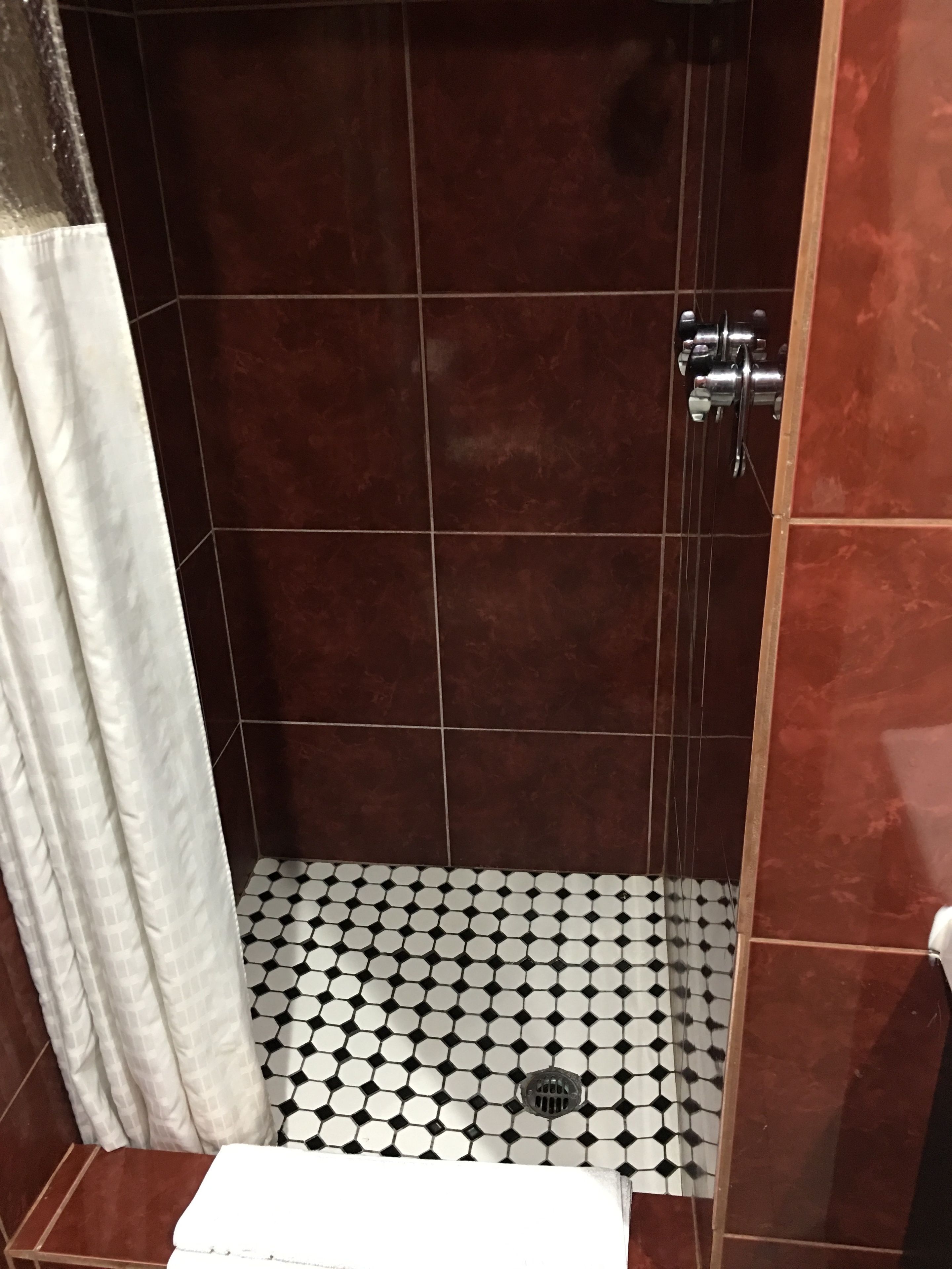 Um chuveiro com azulejos marrons escuros, uma cortina branca e um chão com padrão preto e branco.
