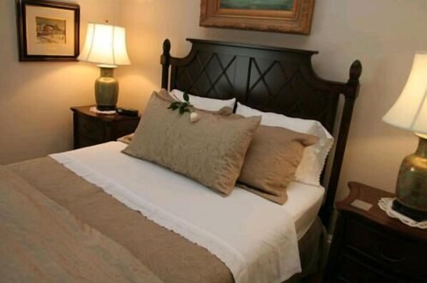 Una cama bien tendida con una colcha blanca y dos almohadas color beige, un cabecero de madera y dos lámparas de noche sobre mesitas de noche de madera.