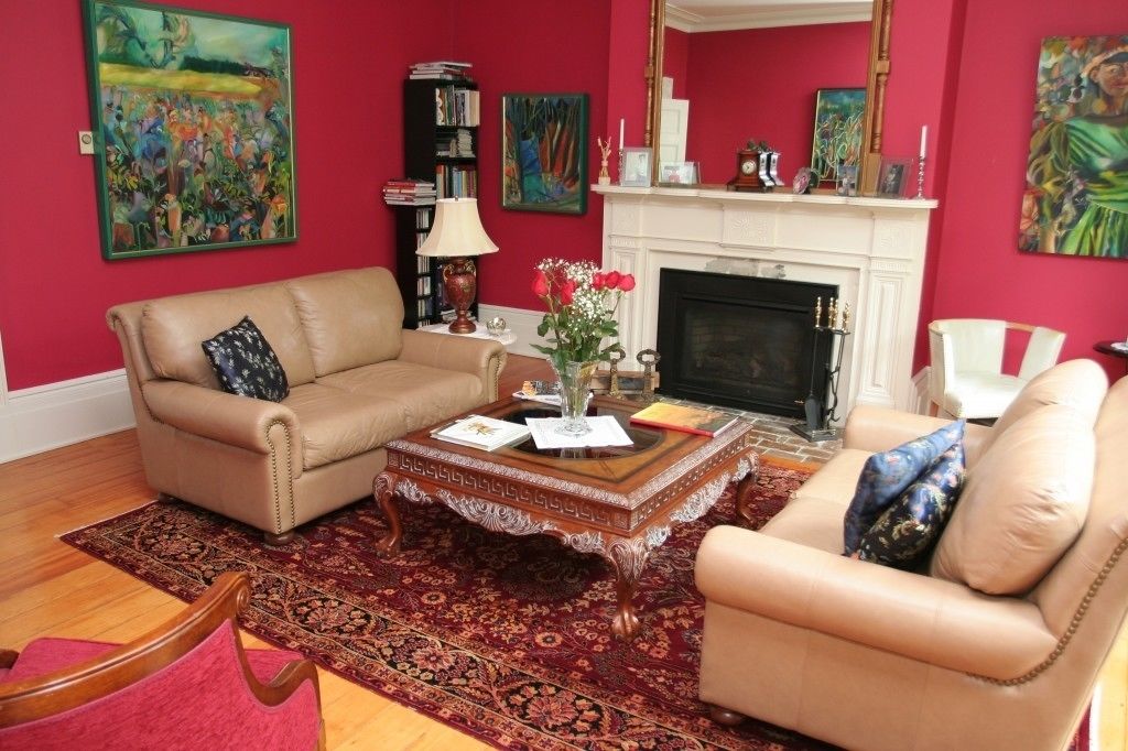 Ein Wohnzimmer mit einem beigefarbenen Sofa, einem passenden Sessel, einem Holztisch und einem roten Teppich mit Muster. Ein Kamin, ein Bücherregal und ein Gemälde an der Wand sind ebenfalls zu sehen.