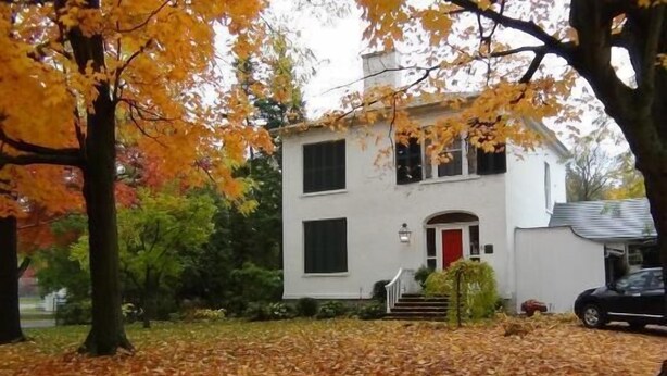 A two-story white house with a red door, surrounded by trees with autumn leaves. - Historic Lyons House (Niagara-on-the-Lake)