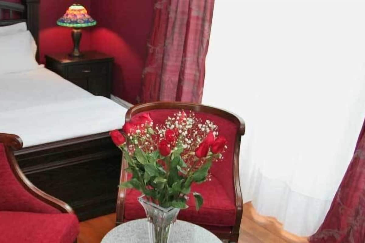 A neatly made bed with white linens, a red upholstered chair, a small round table with a vase of flowers, and two bedside lamps.