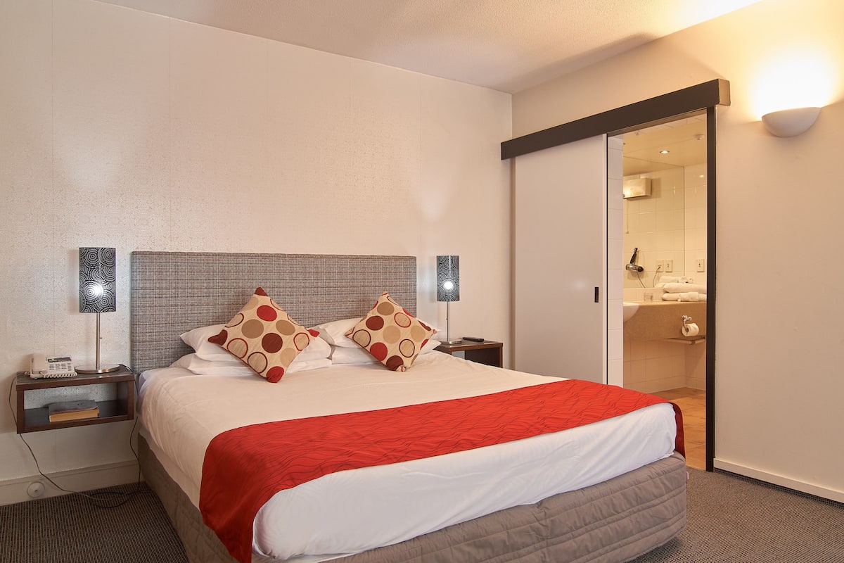 A neatly made bed with a red blanket, two bedside lamps, and a nightstand with a phone and books.