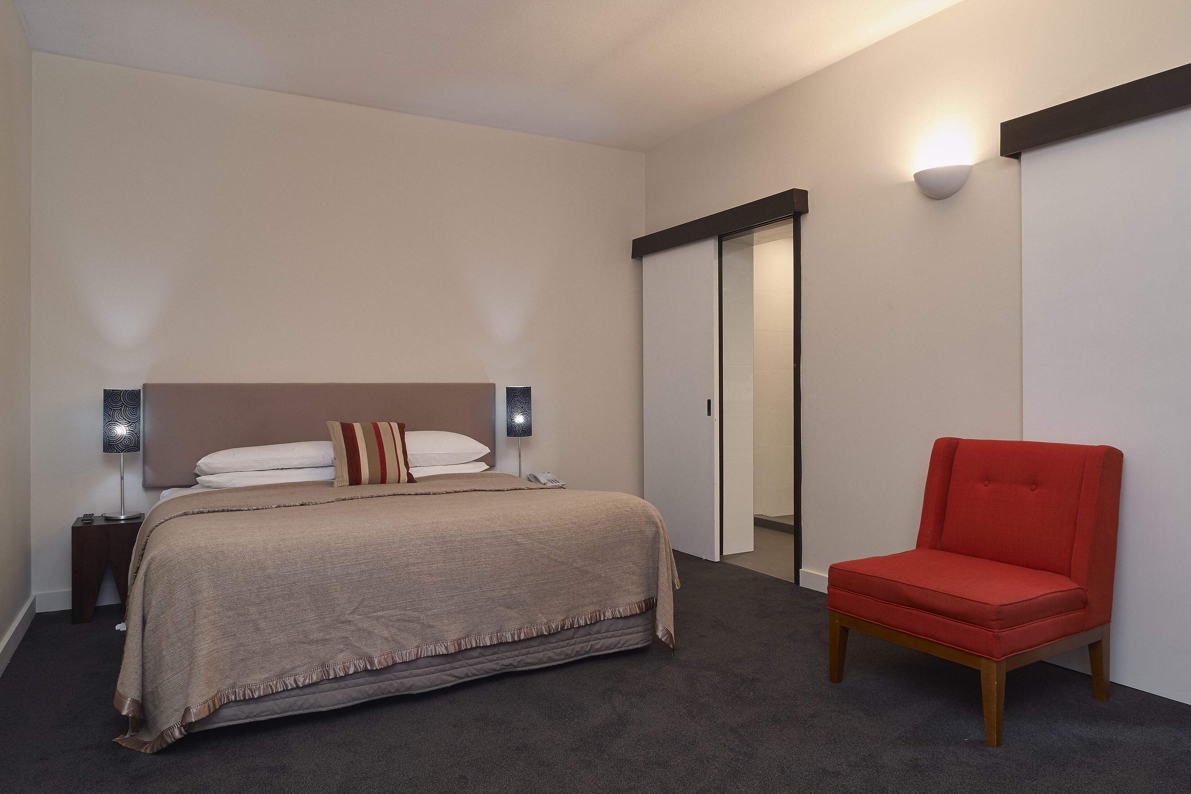 A bedroom with a bed, a red chair, a wardrobe, and a nightstand with a lamp.