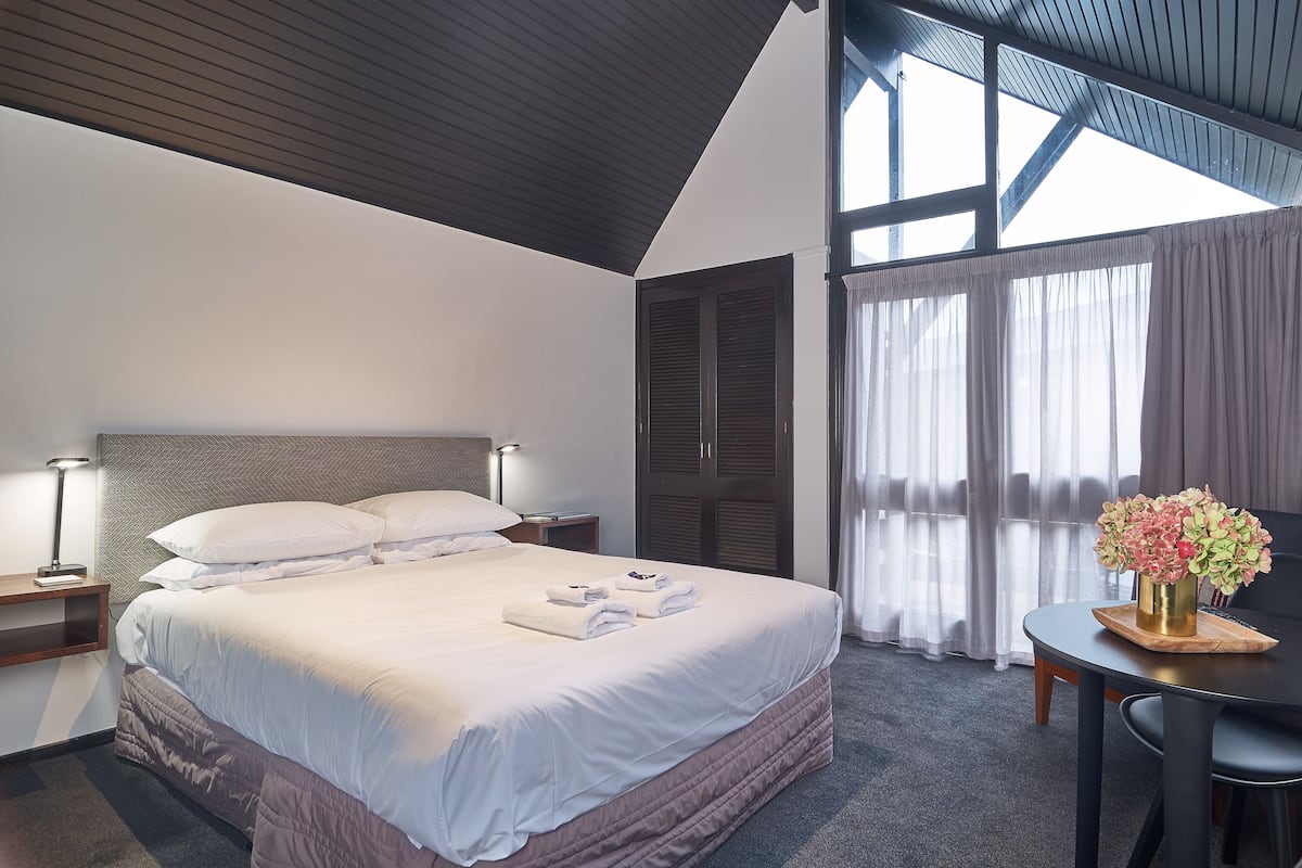 A neatly made bed with white linens, a bedside table with a lamp, and a small table with a vase of flowers.