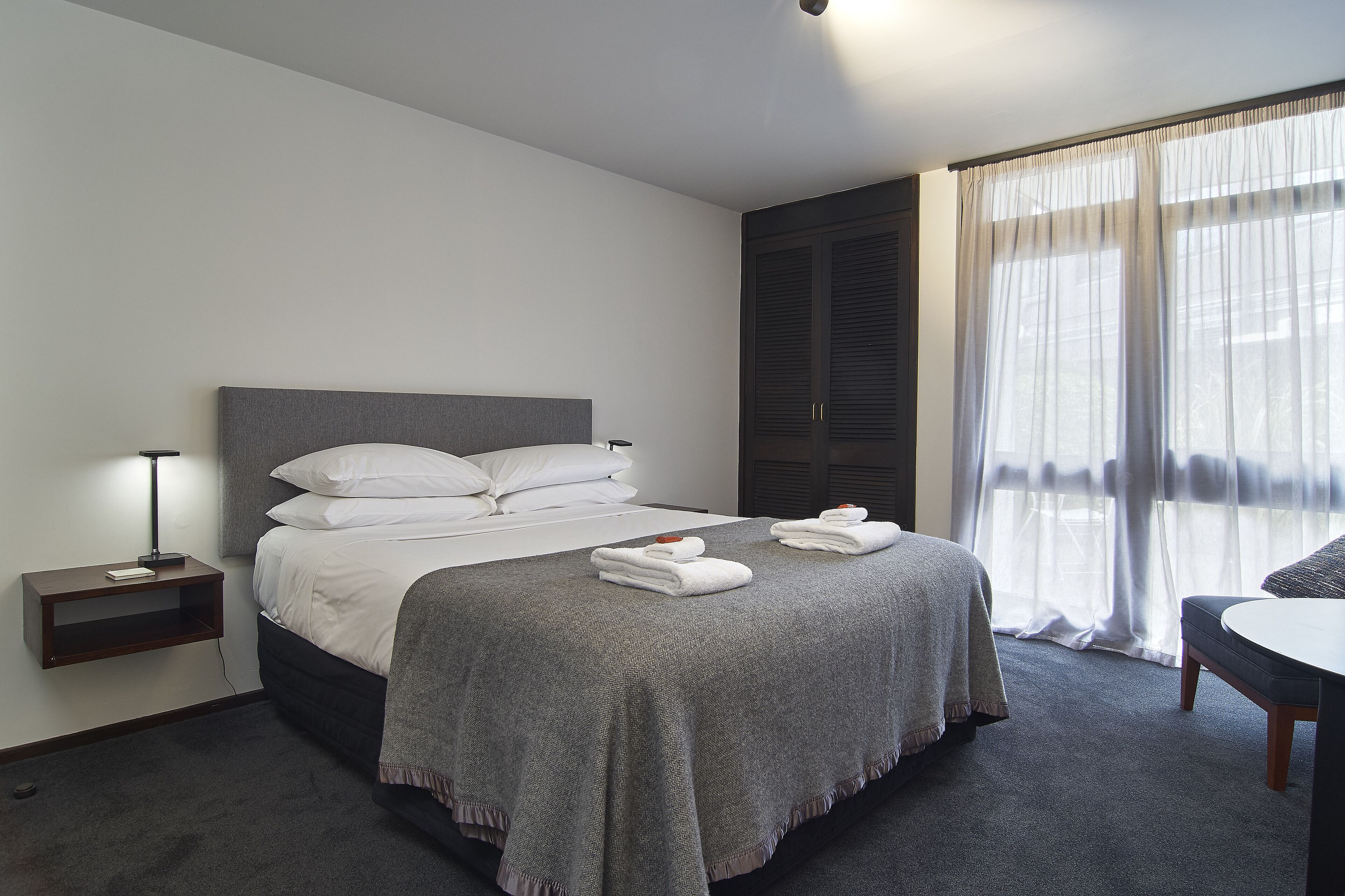 A neatly made bed with white linens and a grey blanket, a bedside table with a lamp, and a chair with a table.