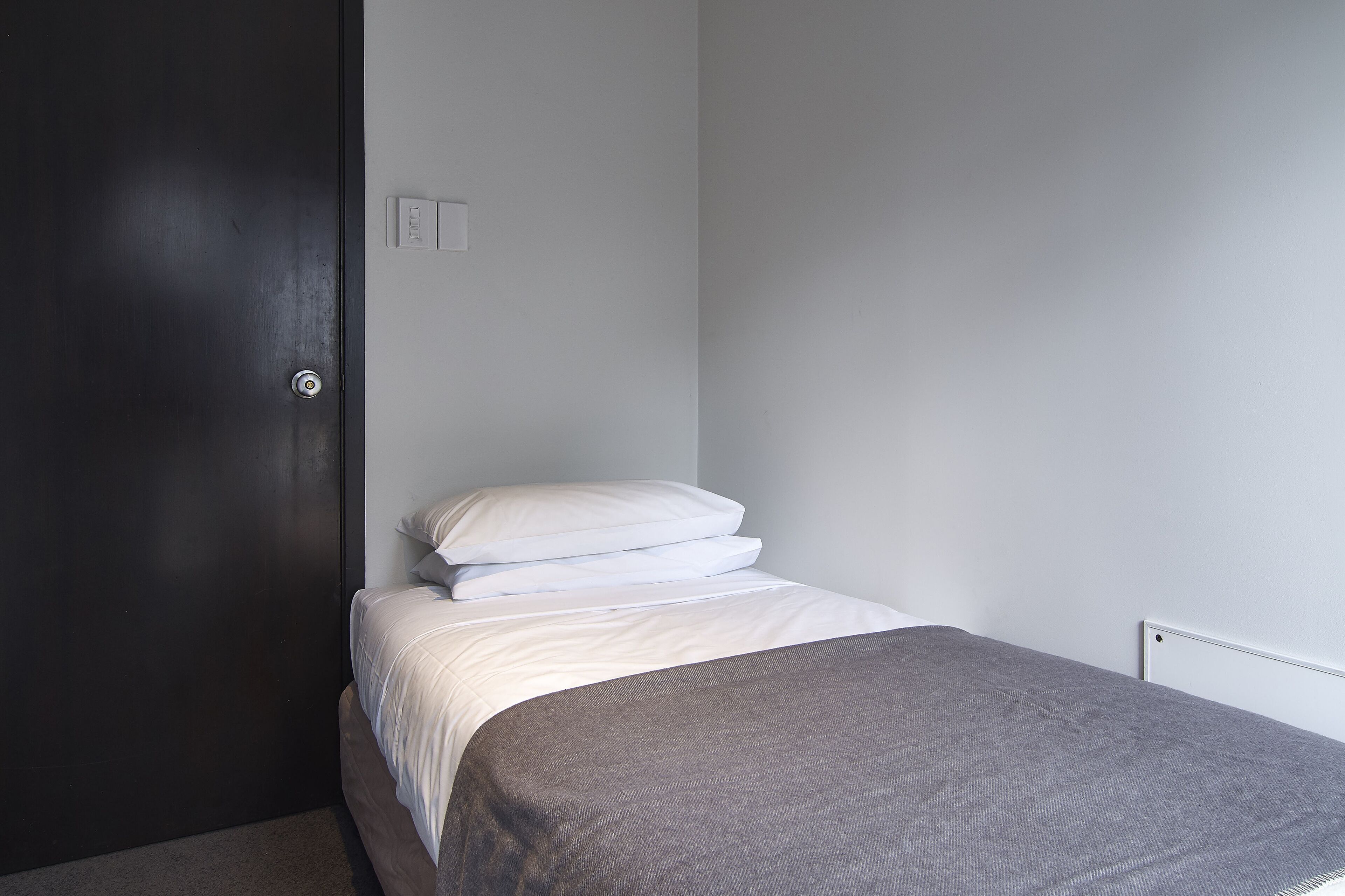 A small, neatly made bed with white bedding and a gray blanket, a dark door, and a white wall with a light switch.