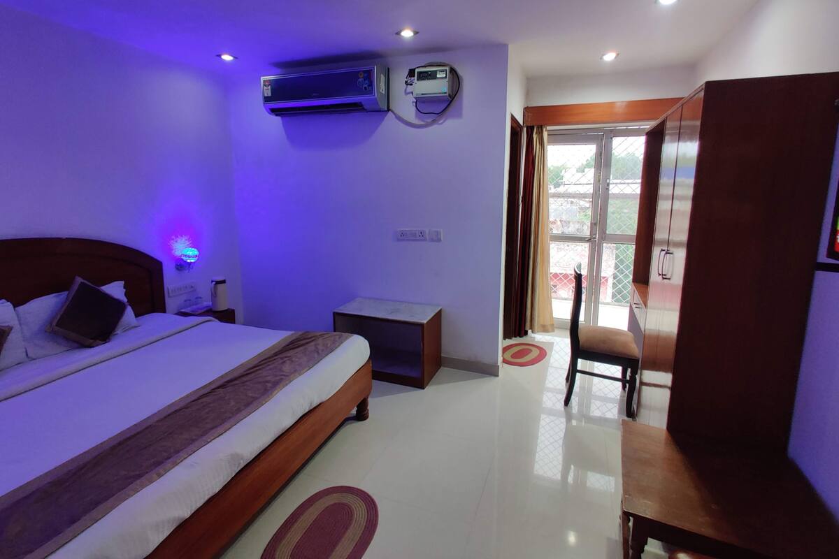 A hotel room with a bed, bedside table, air conditioning unit, and a view of the outside.