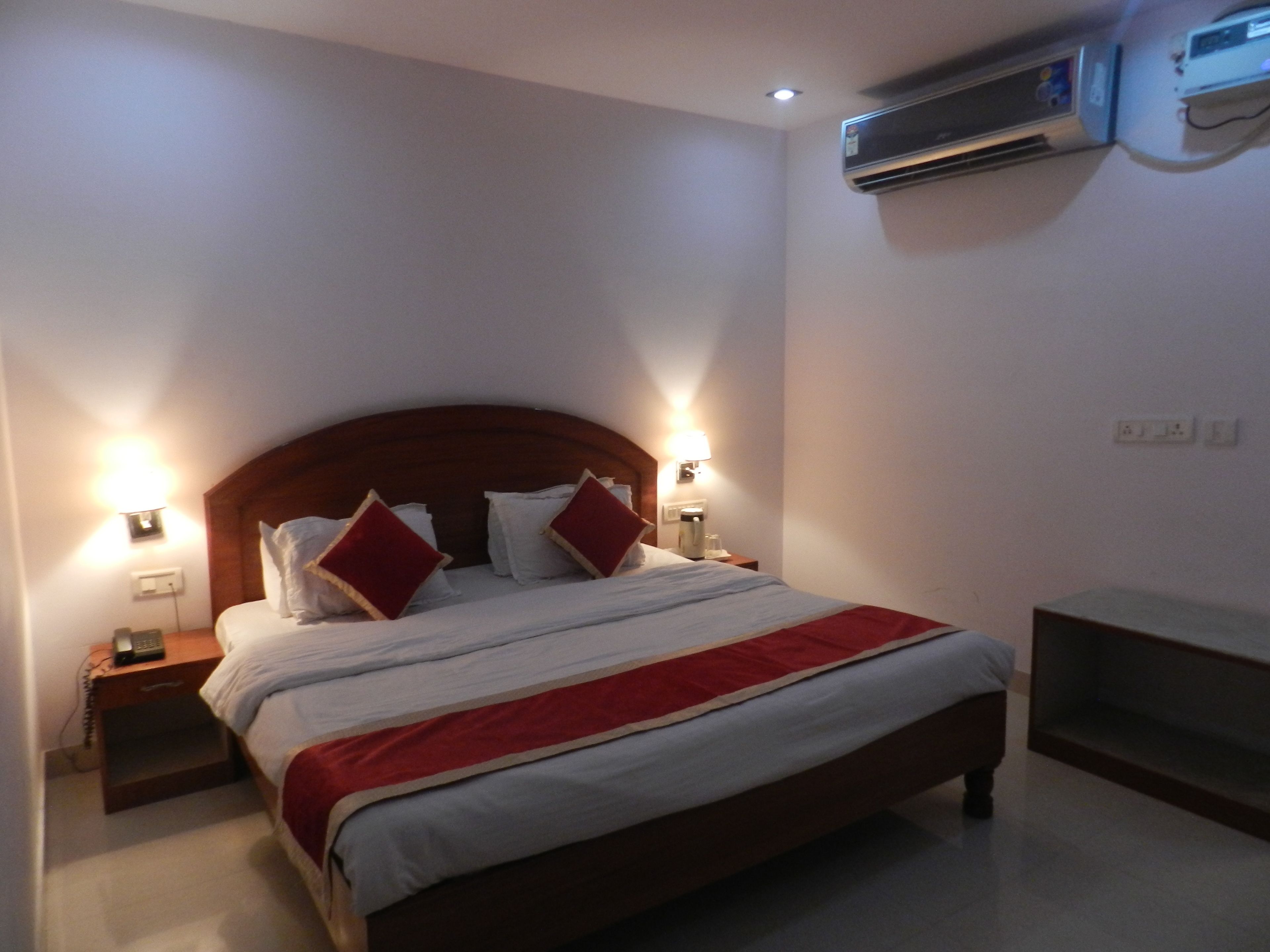 Une chambre d’hôtel avec un lit en bois, deux tables de chevet, une petite table d’appoint, une couvre-lit rouge et blanche et un climatiseur fixé au mur.