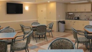A dining area with tables and chairs, a television mounted on the wall, and a kitchenette with cabinets and appliances.