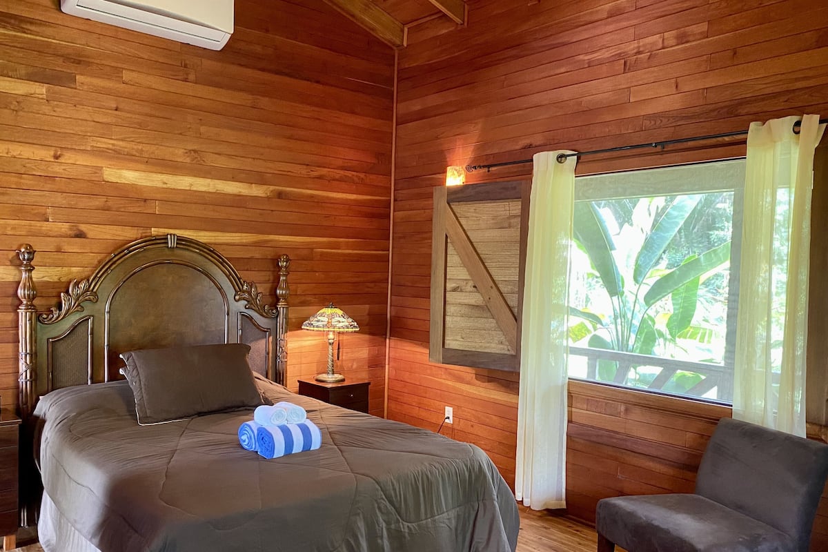 A wooden-paneled room with a bed, a nightstand, a lamp, a chair, and a window with curtains.