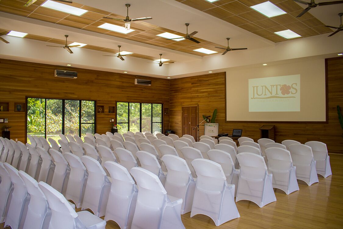 Una sala de conferencias con filas de sillas blancas, paneles de madera, ventiladores de techo y una pantalla de proyector que muestra 