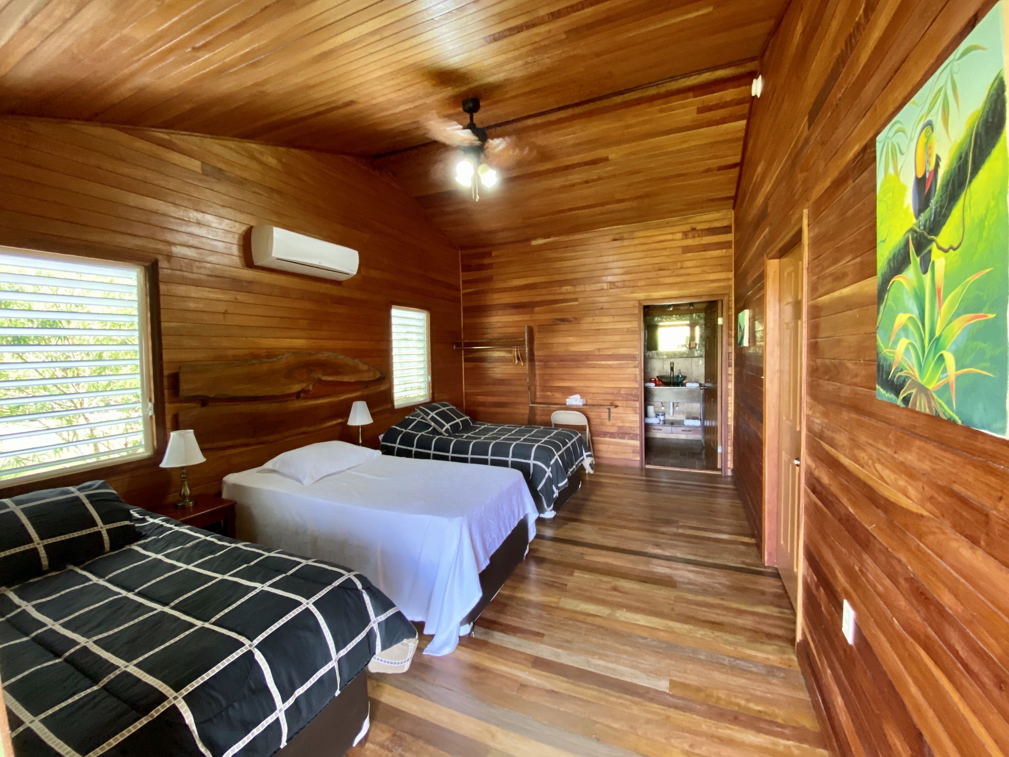 Una cabaña con paredes y suelos de madera, dos camas con ropa de cama a cuadros, un ventilador de techo y un cuadro de un tucán.