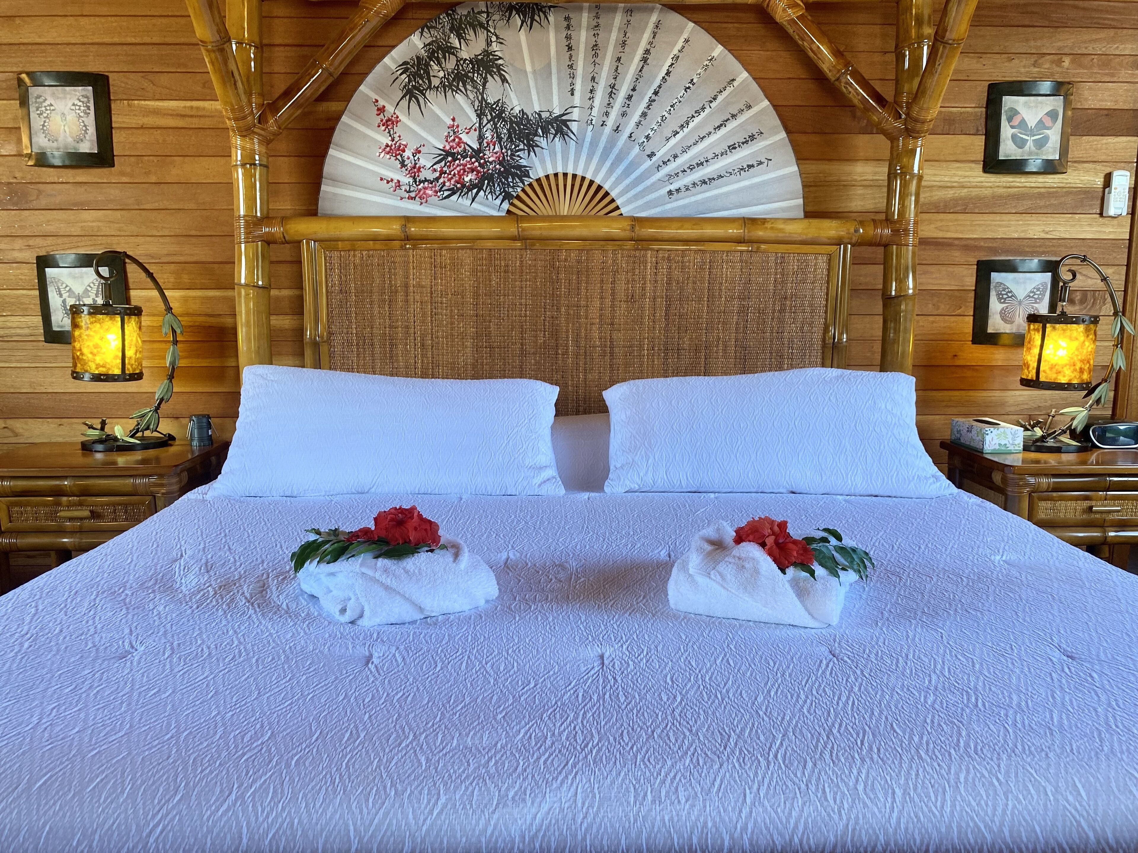 A neatly made bed with white linens and two red flowers on the pillows, a bamboo headboard, and a traditional fan with calligraphy on the wall behind.
