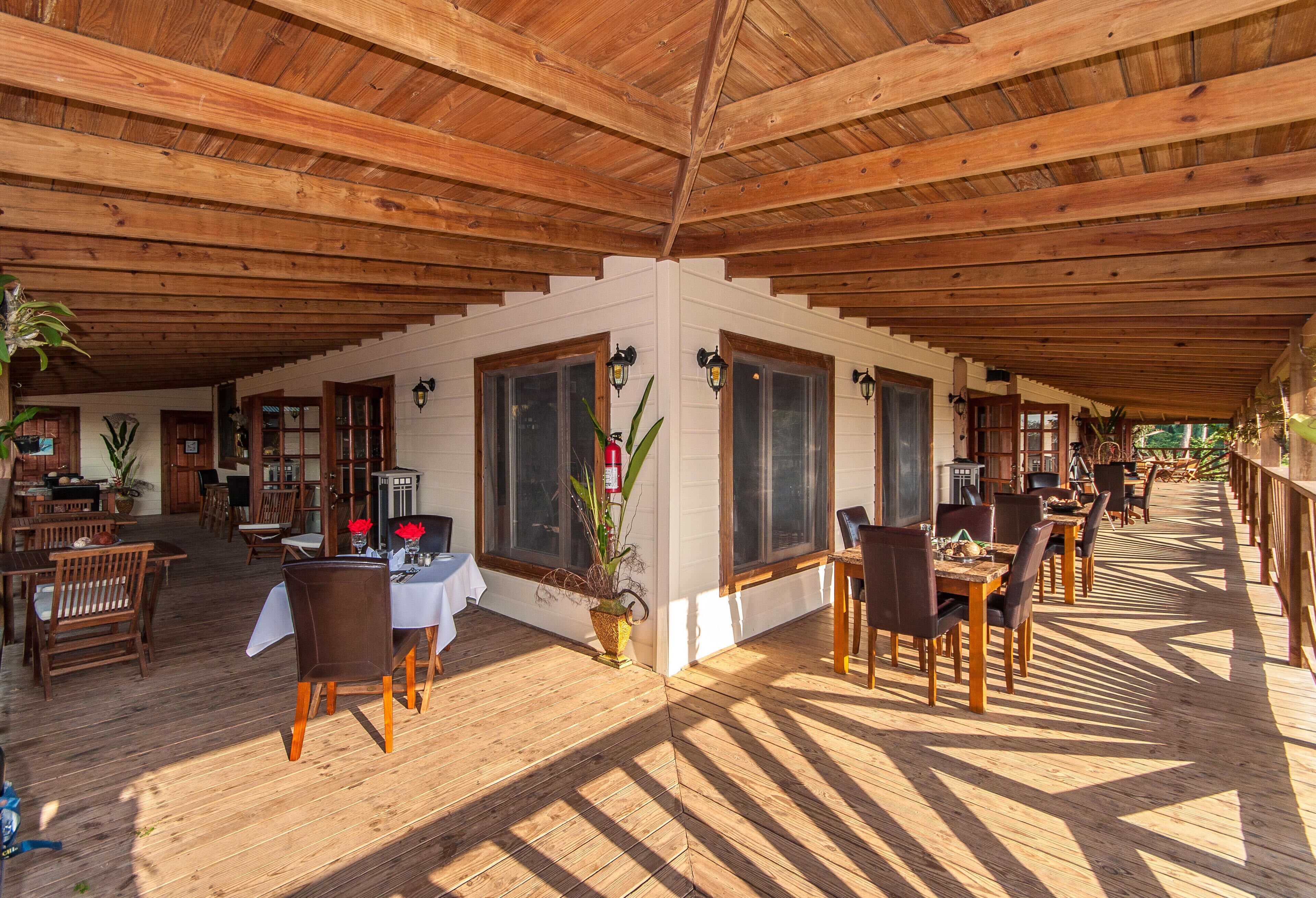 Un espacio exterior para comer con mesas y sillas de madera, ventanas amplias y techo de madera.