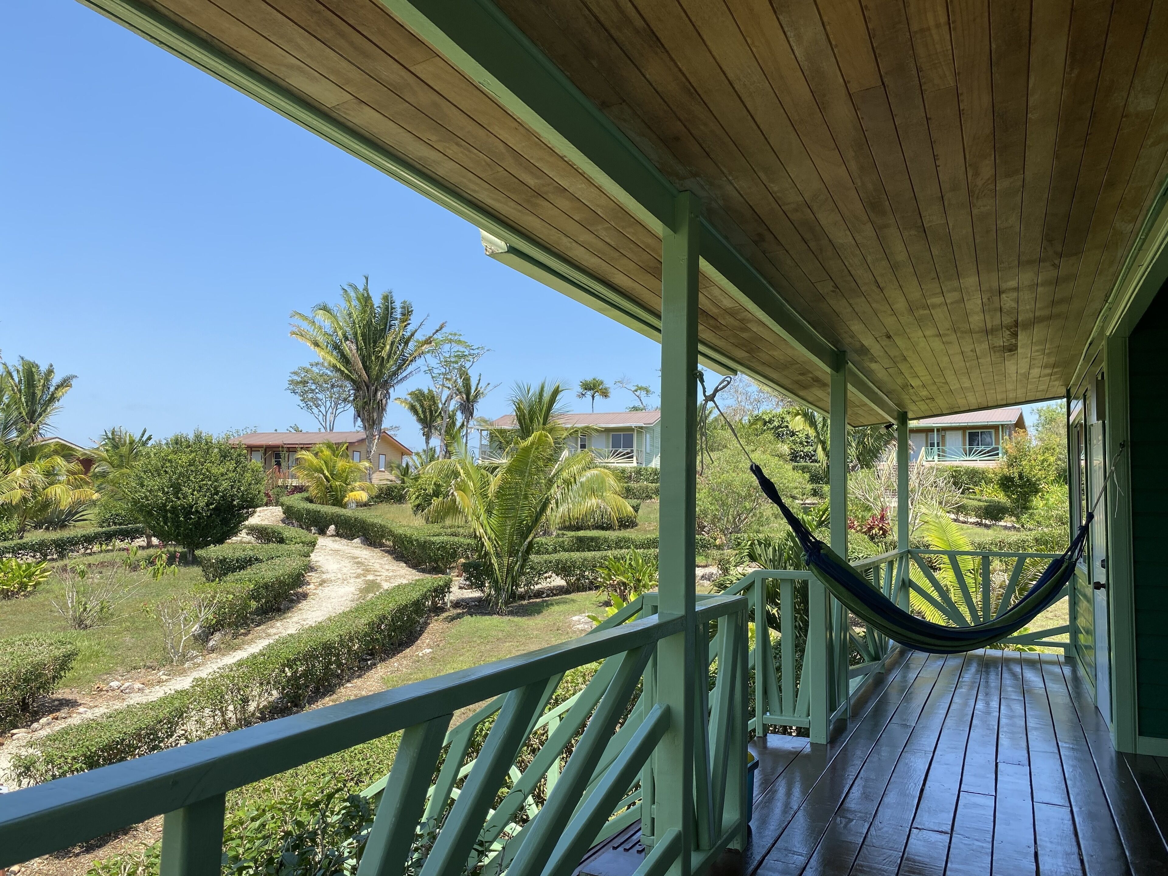 A covered porch with a hammock, a wooden deck, and a view of a landscaped garden with palm trees and a pathway.