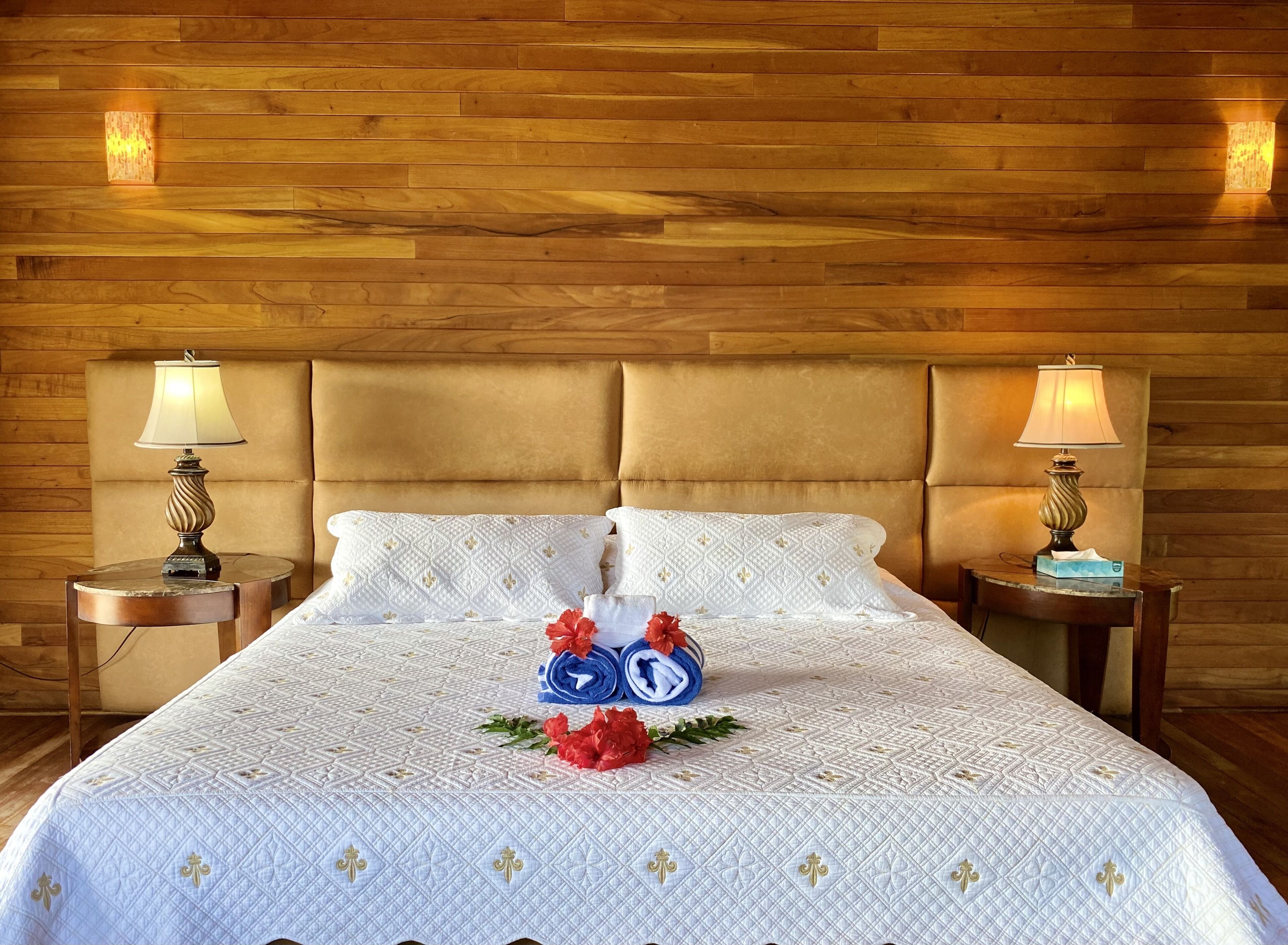 A neatly made bed with a white quilted bedspread, two pillows, and a decorative arrangement of rolled towels and flowers.