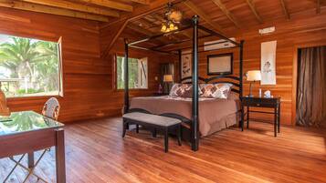Una cama con dosel de madera, acompañada de un banco, una mesa con sillas y una ventana con vista a palmeras.