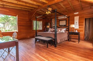 A wooden four-poster bed with a bench in front, a table with chairs, and a window with a view of palm trees. - Dream Valley Belize (Belmopan)