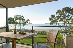 A balcony with a wooden table, two wine glasses, and a bottle of wine. The view includes a beach and trees.