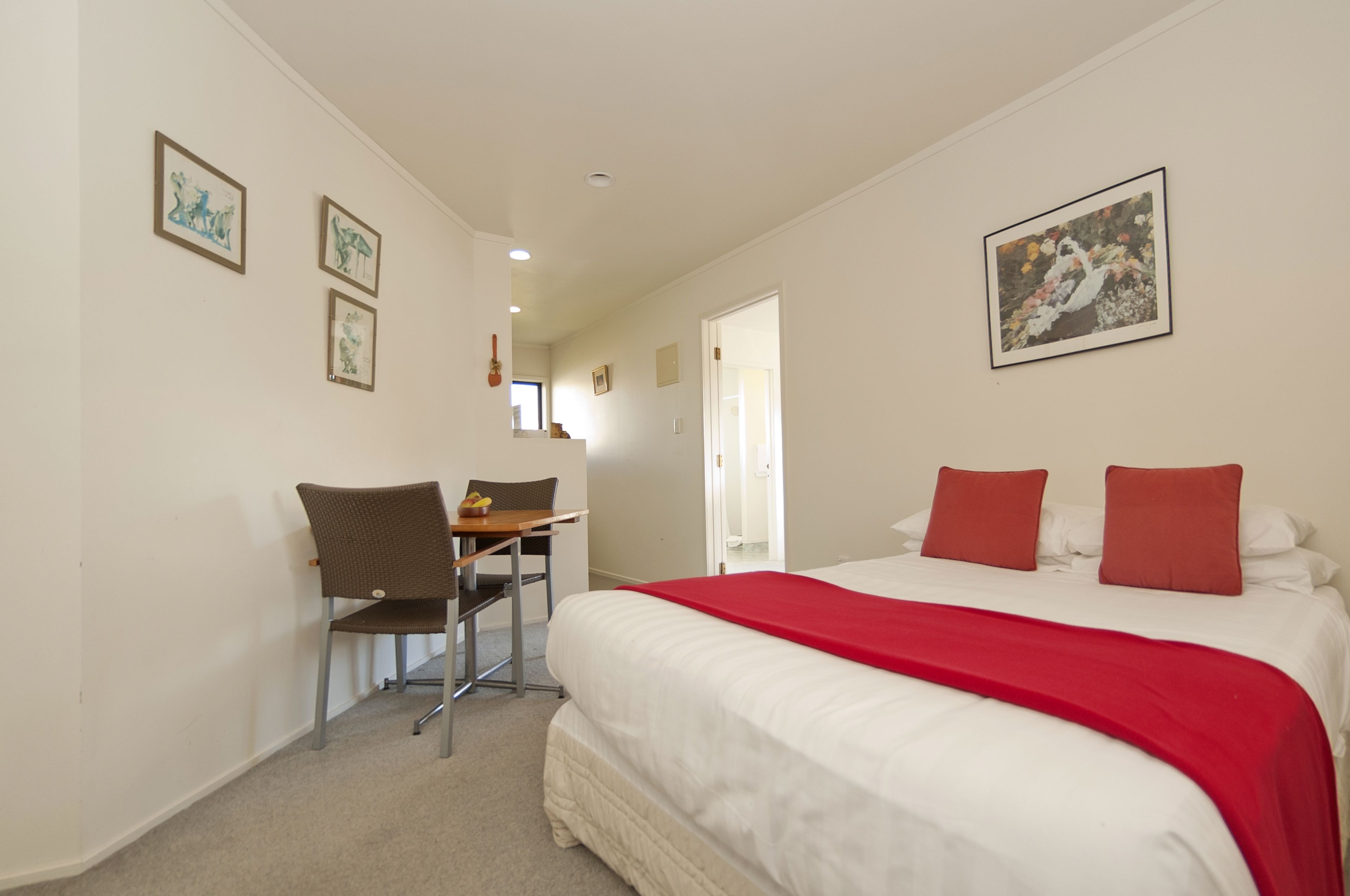 A hotel room with a bed, red pillows, a desk with a chair, and framed artwork on the walls.
