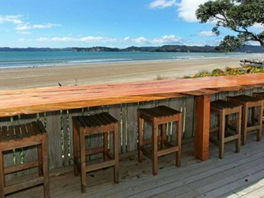 Terraza de madera con un largo banco y taburetes, con vistas a una playa, cielos despejados y colinas a lo lejos.