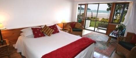 A bedroom with a bed, pillows, a nightstand, a chair, a table, and a view of the beach through large windows.
