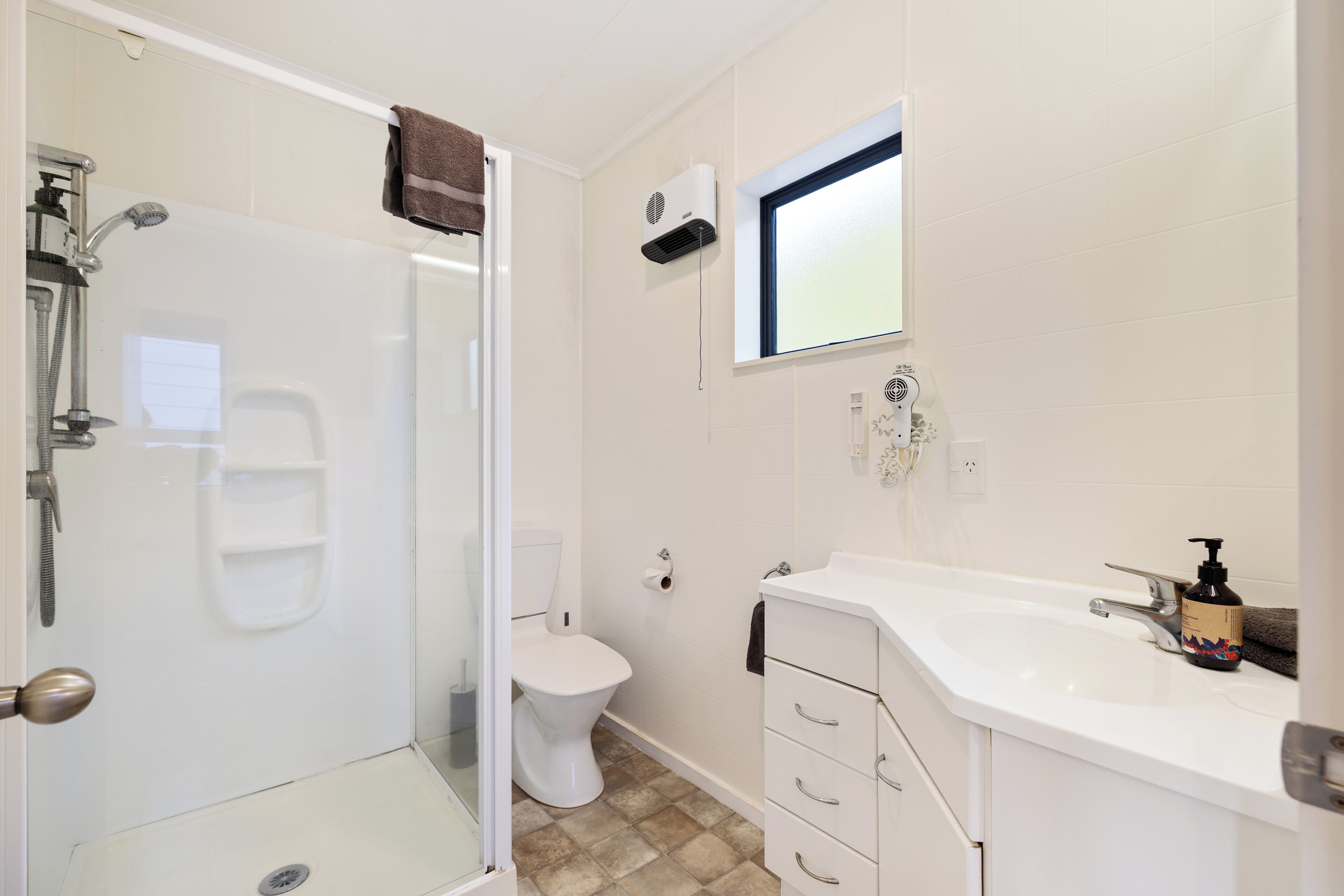 A bathroom with a glass-enclosed shower, a toilet, a sink with a soap dispenser, and a window above the sink.