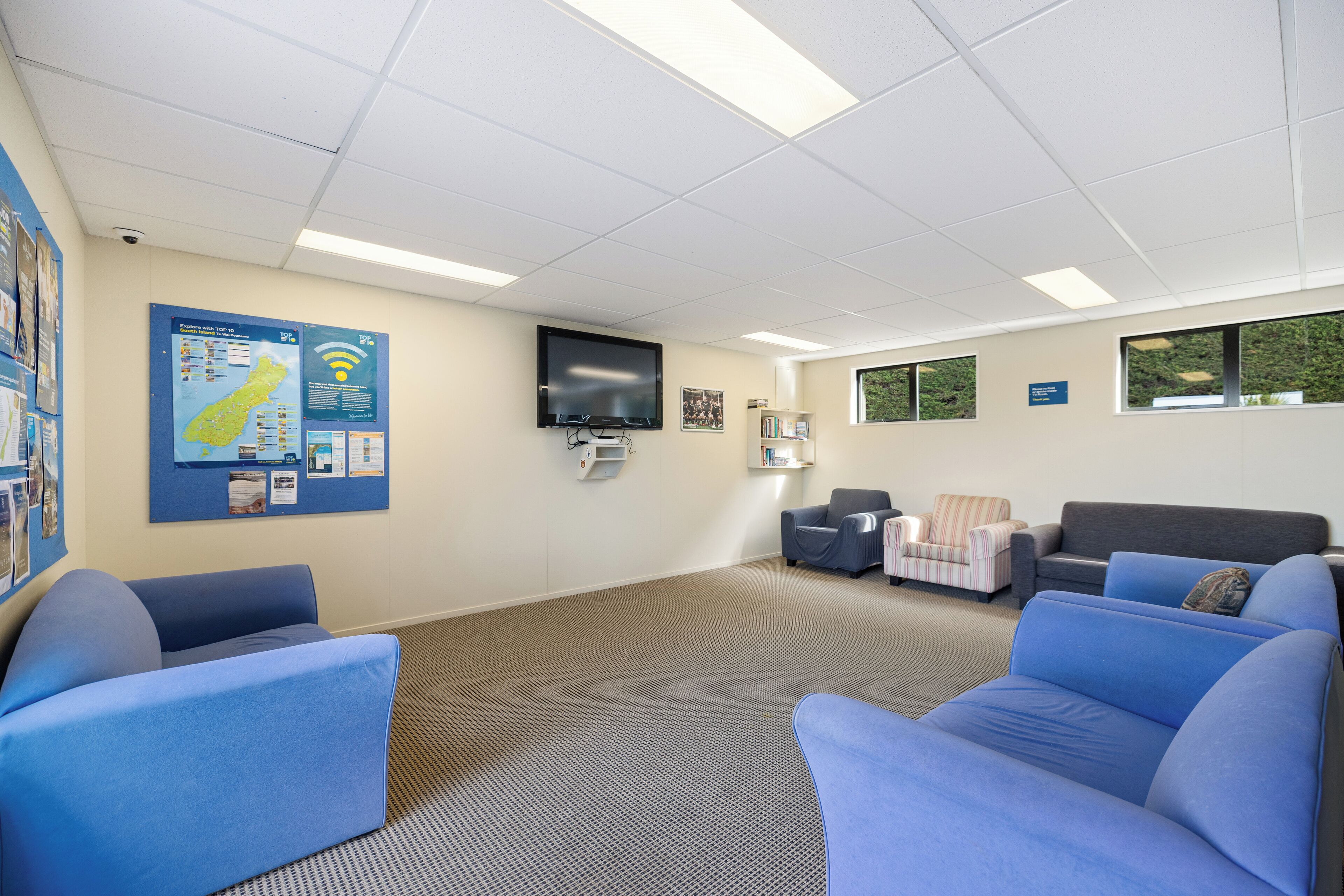 Une salle d’attente avec des canapés bleus, un téléviseur à écran plat, un tableau d’affichage et une fenêtre donnant sur un espace verdoyant.